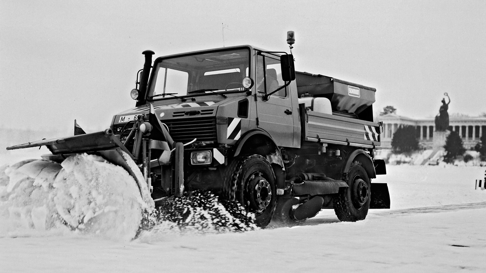 Een Mercedes-Benz Unimog ruimt sneeuw met een sneeuwschuif. De scène toont de inzetbaarheid van het voertuig onder winterse omstandigheden op een met sneeuw bedekte ondergrond.