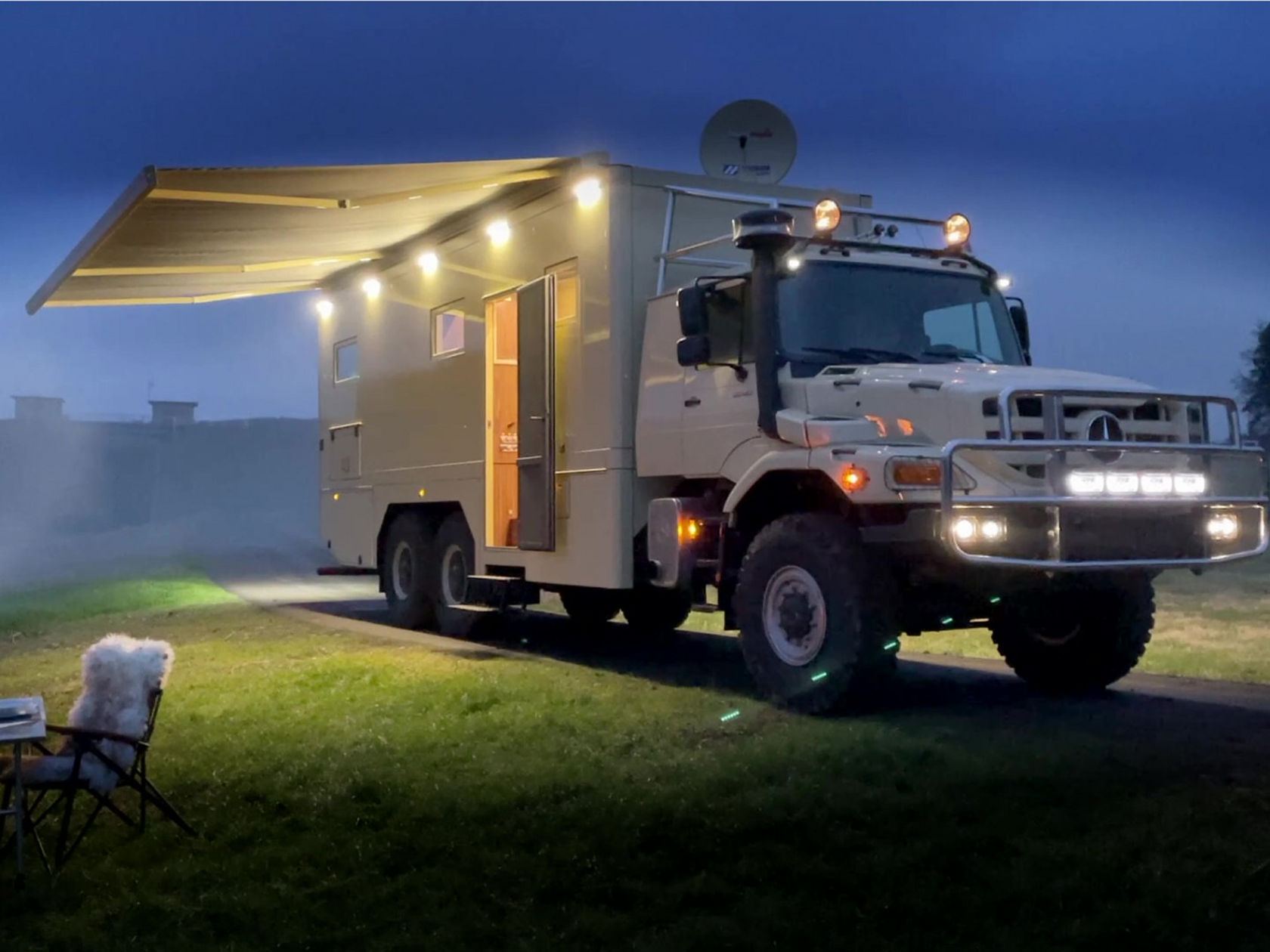 A Zetros as an off-road campervan stands on a small path next to a lawn. It’s evening or night. An awning on the truck is extended and illuminated. A camping chair and table are on the left-hand side of the picture.