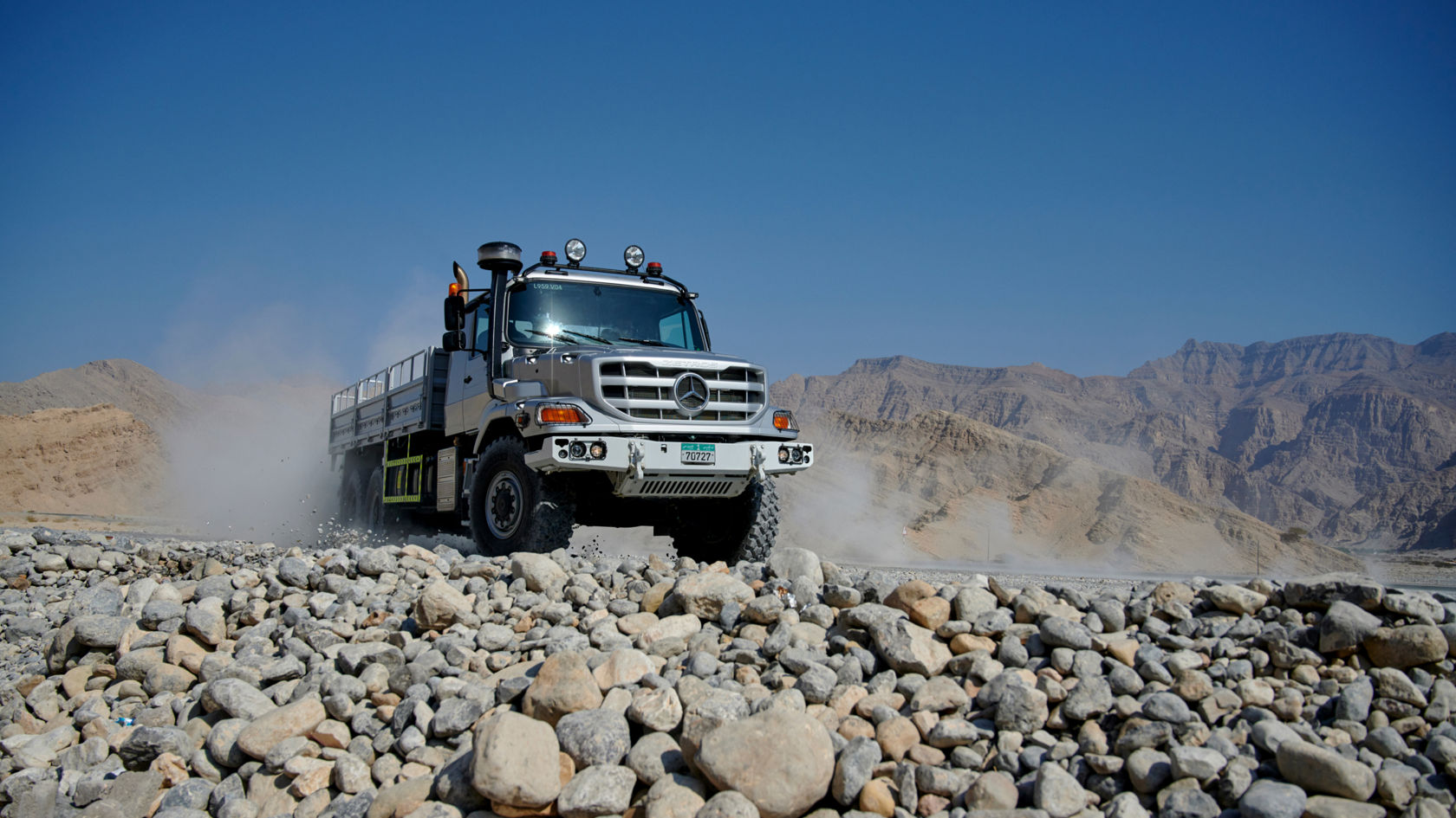 A silver Zetros drives over rocky terrain and swirls up dust. Mountains and blue sky can be seen in the background.