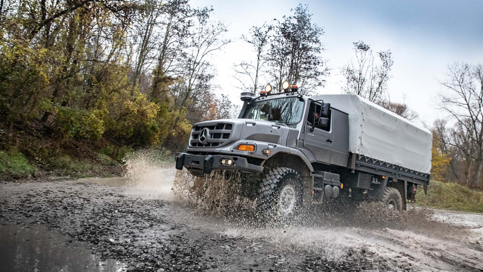 An anthracite grey Zetros forcefully drives through a muddy puddle and splashes water. A tarpaulin is stretched over the cargo area. A forest can be seen in the background.