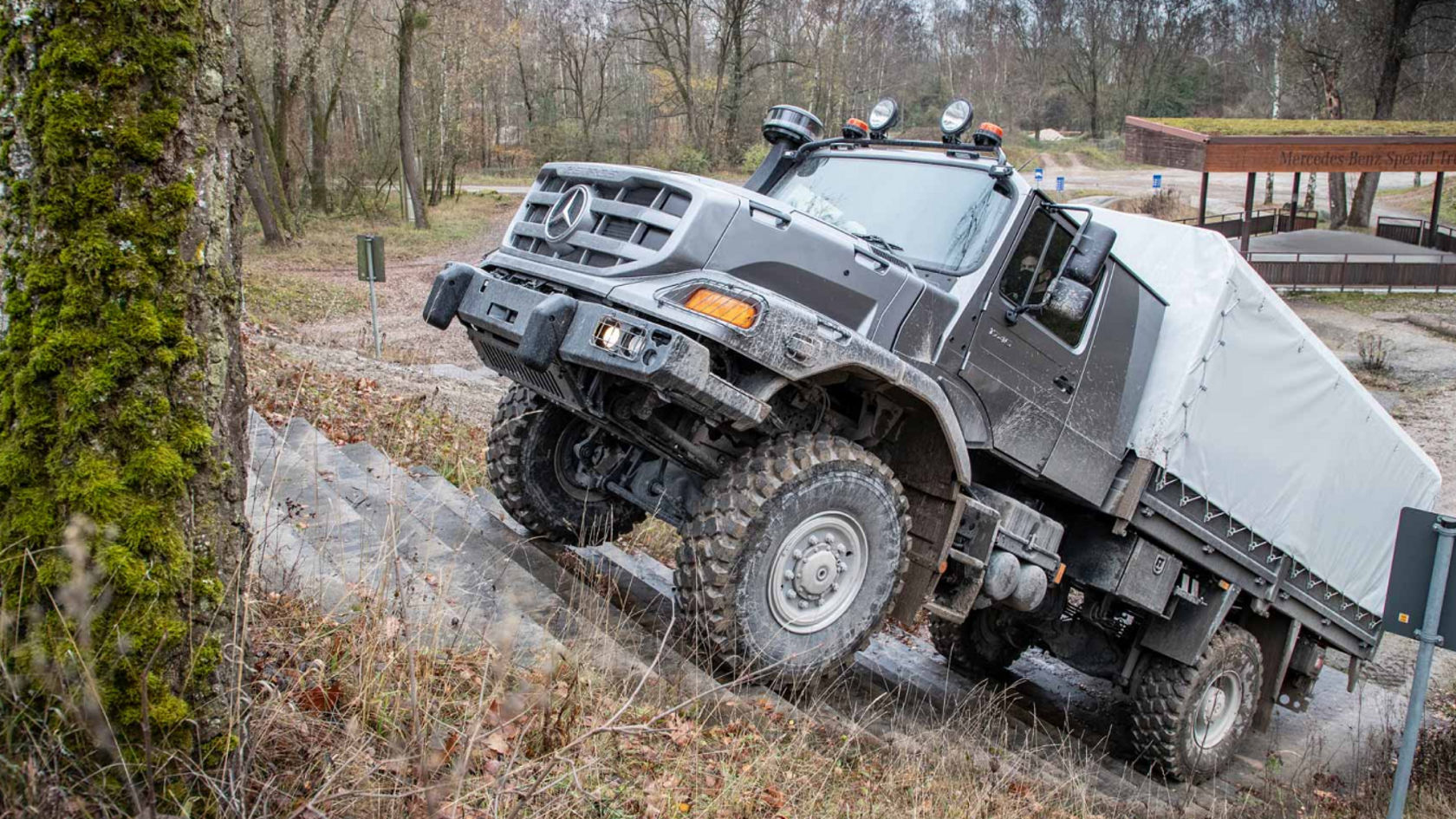 An anthracite grey Zetros drives up a staircase on a test site. Unpaved roads and forests can be seen in the surrounding area.