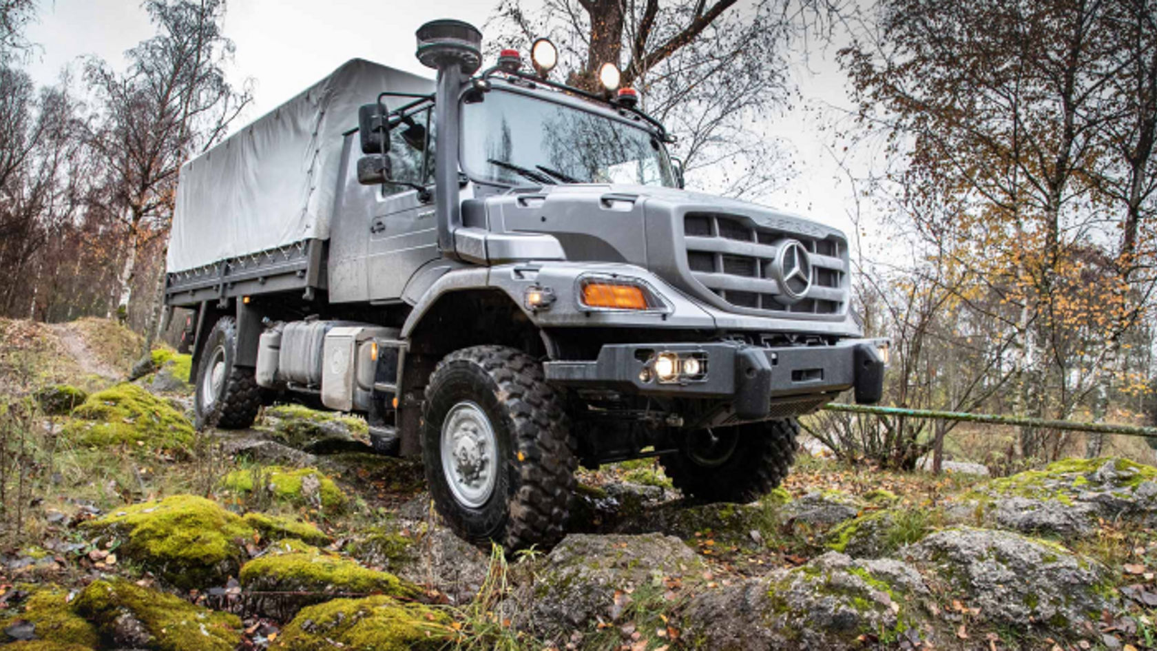 An anthracite grey Zetros with tarpaulin drives over rugged, mossy terrain. Trees and part of a railing can be seen in the background.
