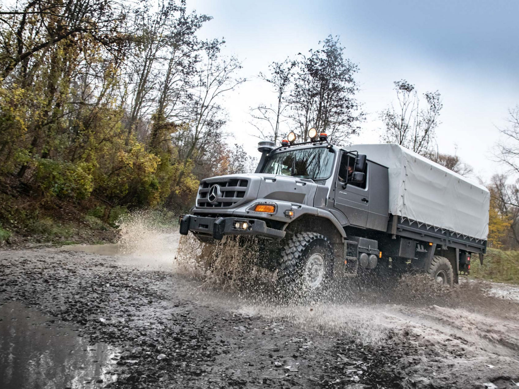 An anthracite grey Zetros forcefully drives through a muddy puddle and splashes water. A tarpaulin is stretched over the cargo area. A forest can be seen in the background.